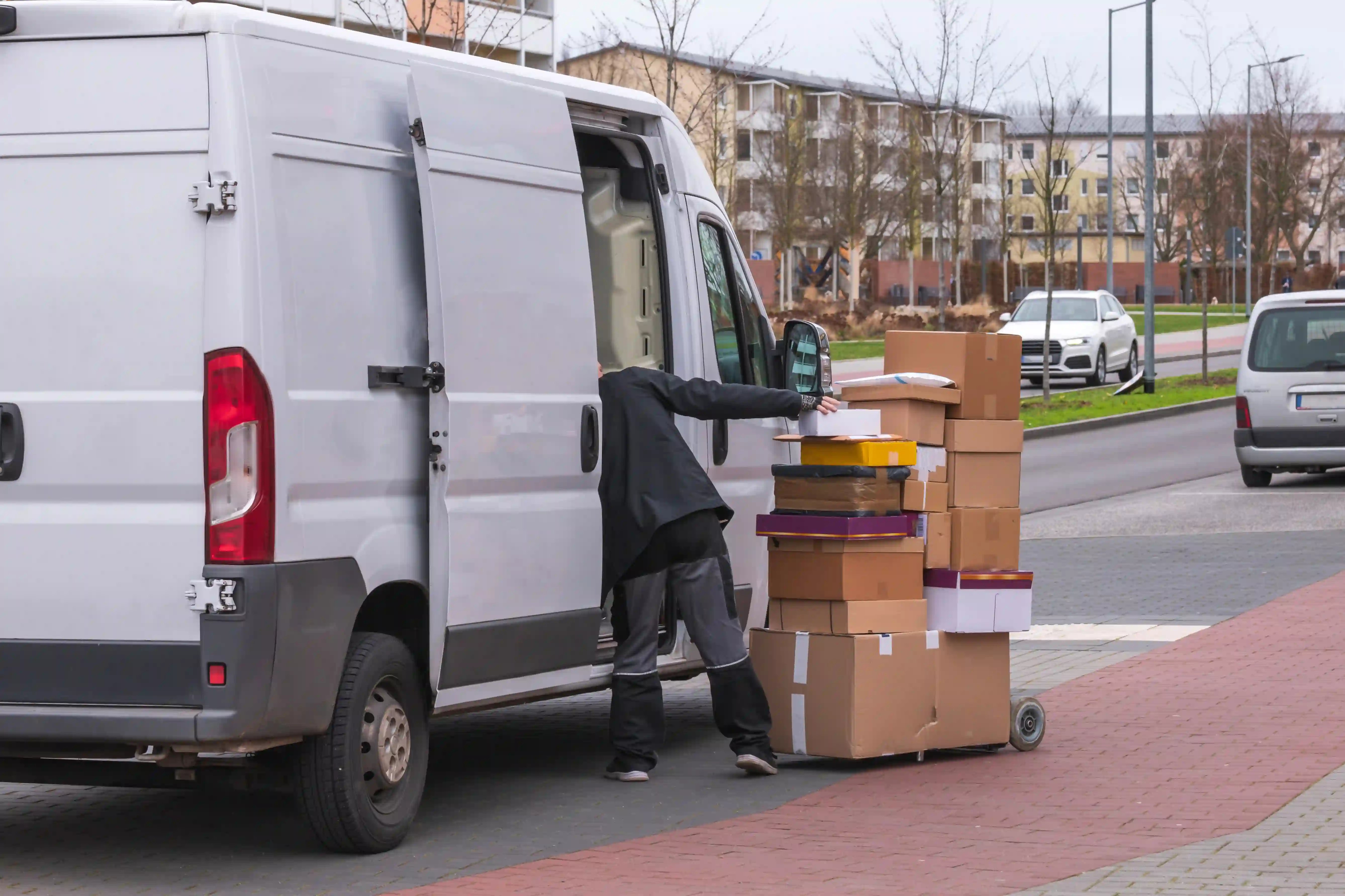 Mann lädt mehrere Kartons von einem Transportwagen in einen weißen Lieferwagen vor einem Wohngebäude.